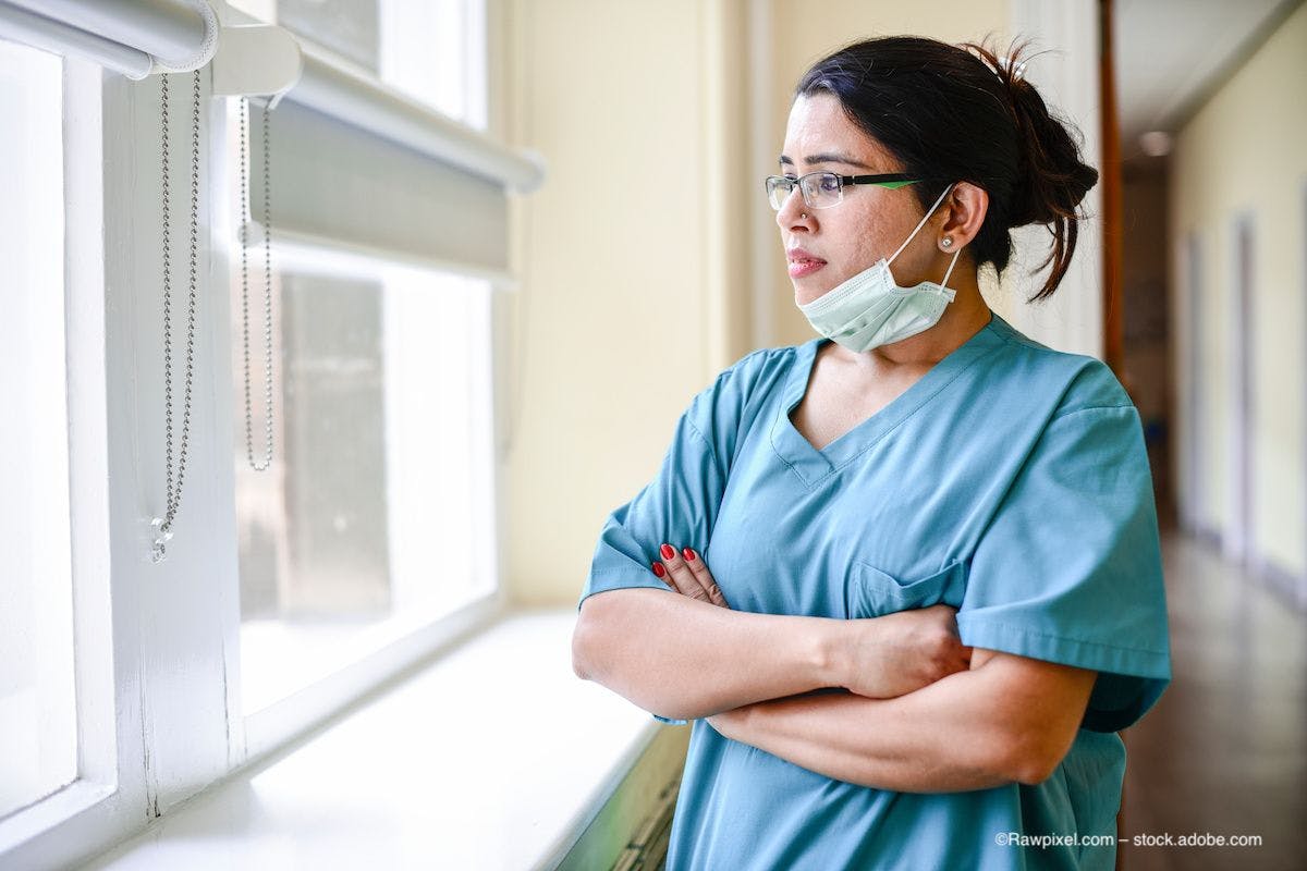 A woman in scrubs stands with her face mask down, looking out the window. Image credit: ©Rawpixel.com – stock.adobe.com