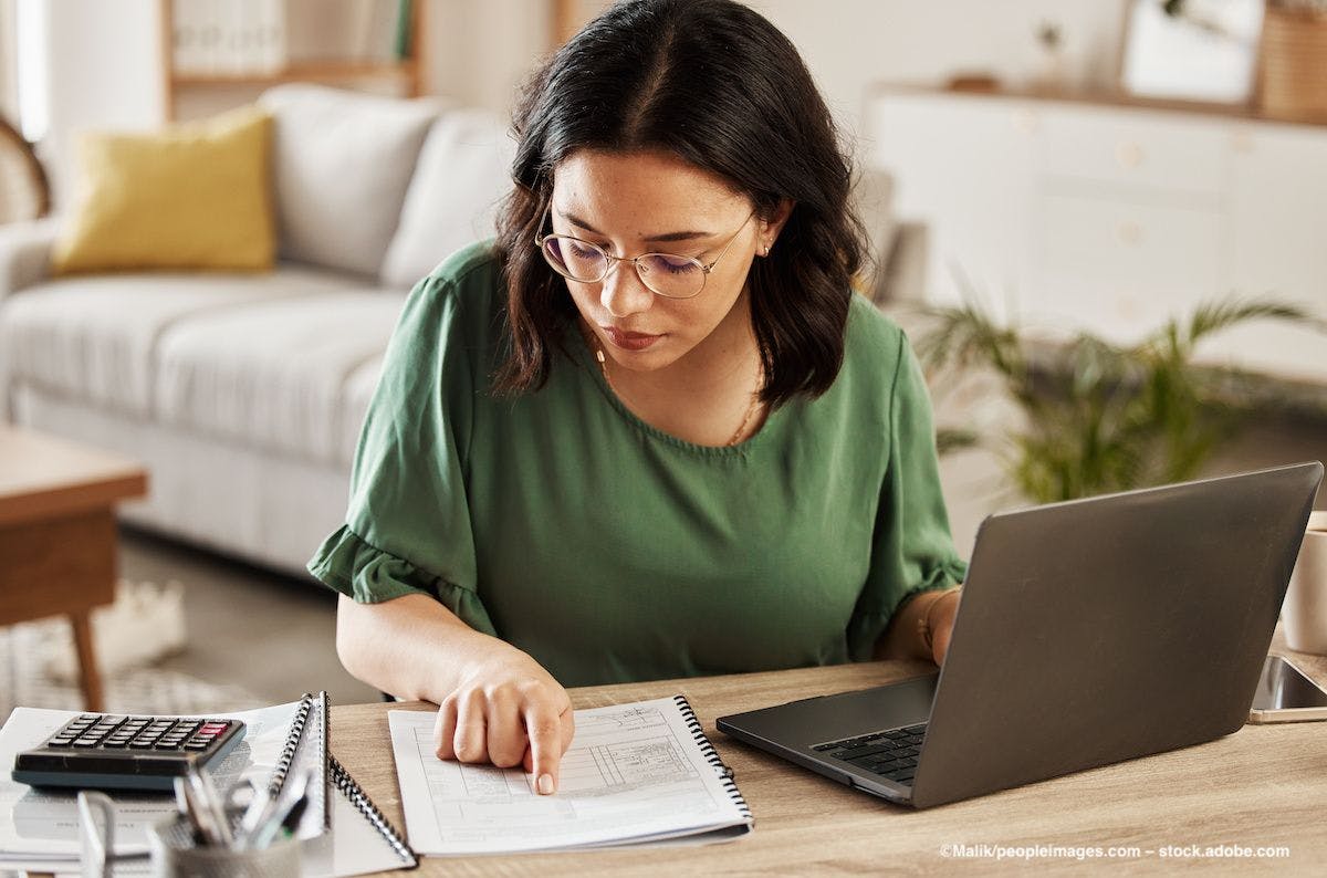 A woman wearing glasses consults paperwork at her desk. Image credit: ©Malik/peopleimages.com – stock.adobe.com