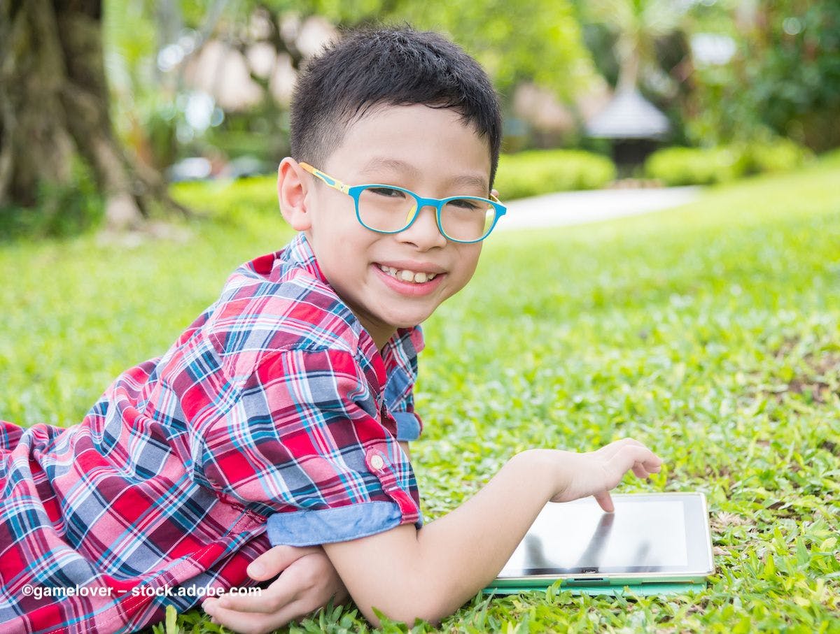 A young boy wearing glasses sits in the grass, playing on a tablet computer. Image credit: ©gamelover – stock.adobe.com