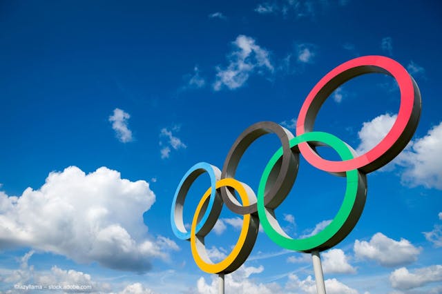 A public art display of Olympic rings stands in front of a blue sky with puffy clouds. Image credit: ©lazyllama – stock.adobe.com