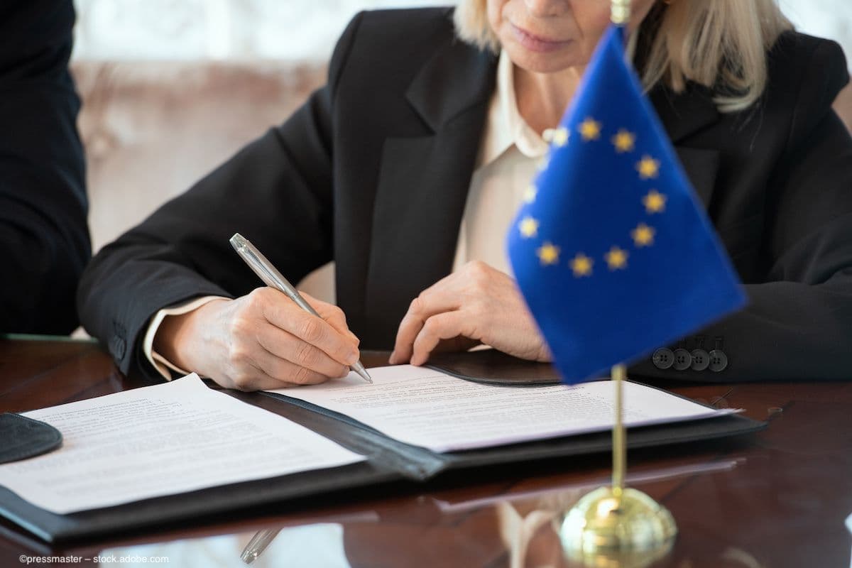 A European Union delegate signs a document while an EU flag is visible on the desk. Image credit: ©pressmaster – stock.adobe.com