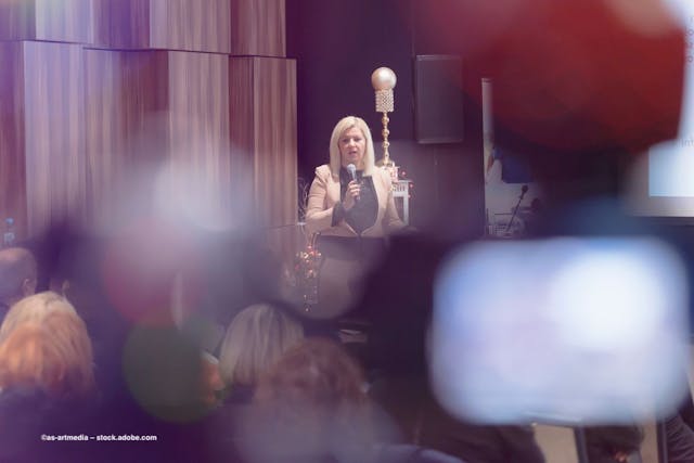 A woman stands at a lectern delivering a presentation to an audience. She is viewed through the blur of the crowded audience. Image credit: ©as-artmedia – stock.adobe.com