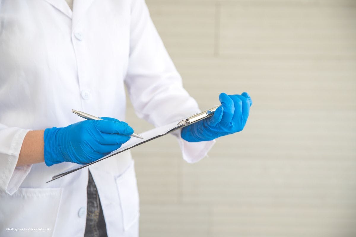 A doctor in a white lab coat and blue surgical gloves holds her clipboard and makes a note. Image credit: ©feeling lucky – stock.adobe.com