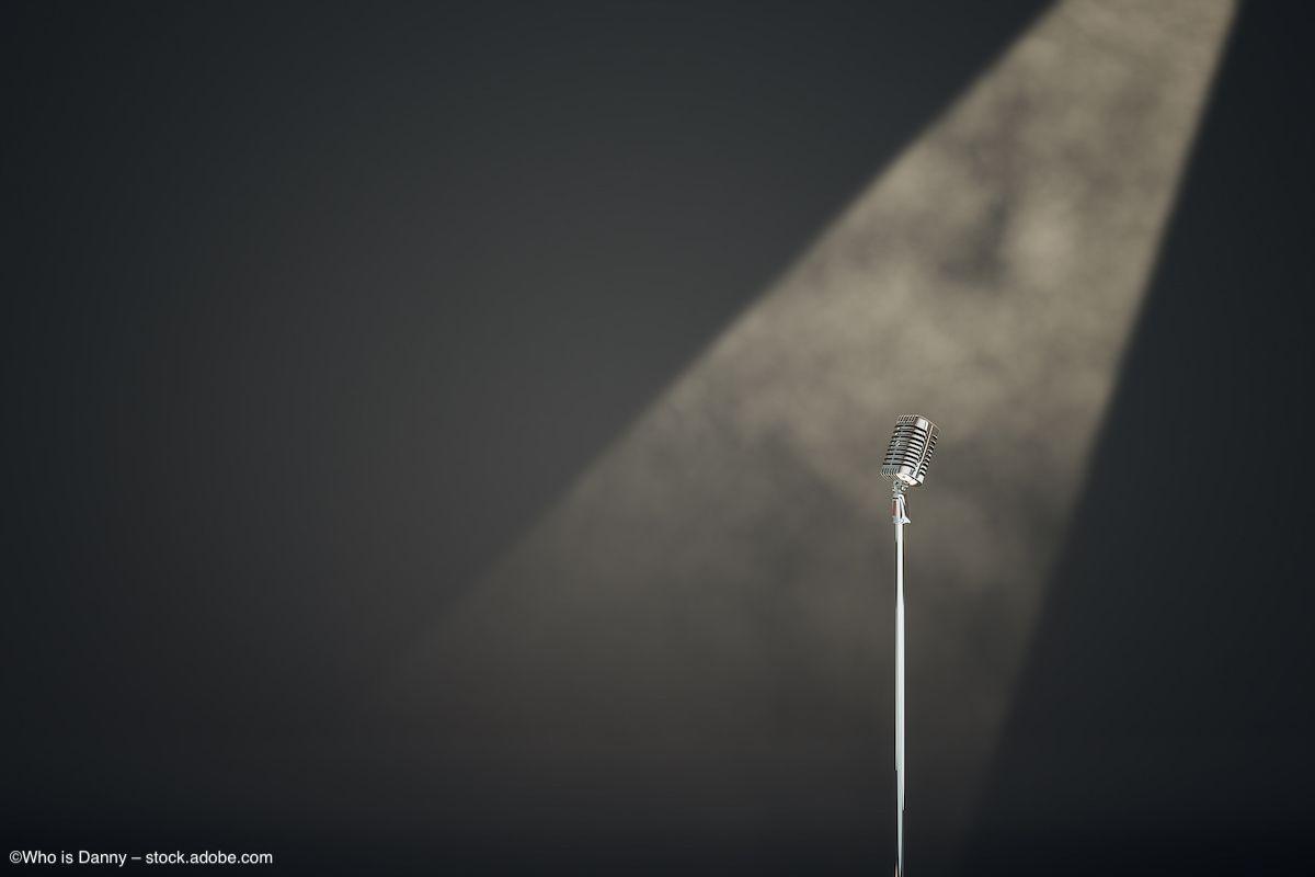 A microphone in a stand is lit by a spotlight, shining on an empty stage. Image credit: ©Who is Danny – stock.adobe.com