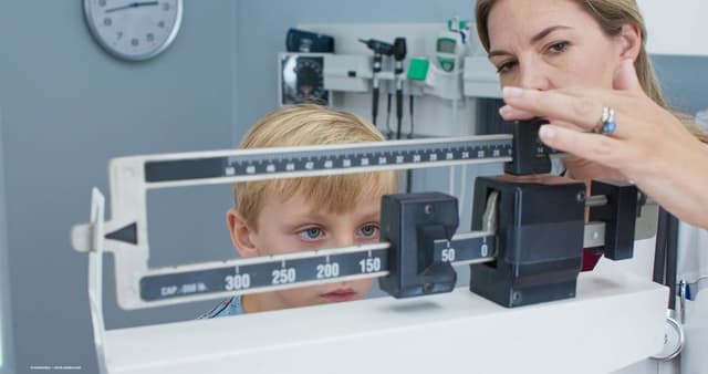 A child stands on a scale in the doctor's office and looks at the scale with the doctor. Image credit: ©rocketclips – stock.adobe.com