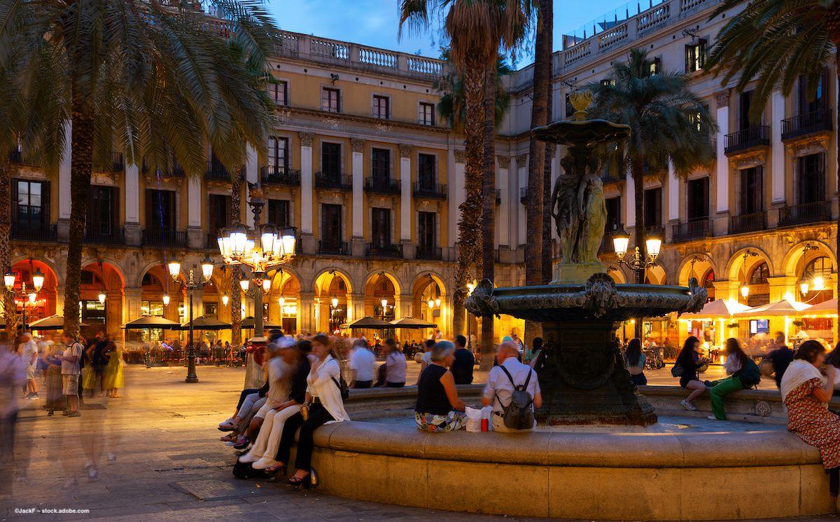 Placa Reial in Barcelona, Spain. A fountain is lit up by streetlamps. Tourists gather at night. Image credit: ©JackF – stock.adobe.com