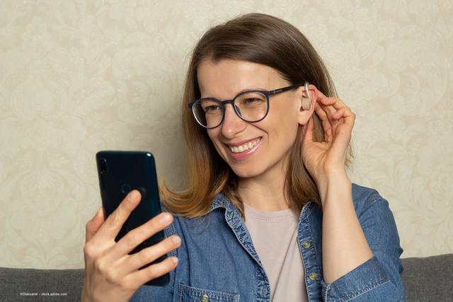 A person with glasses and a hearing aid looks at her phone. Image credit: ©Oleksandr – stock.adobe.com