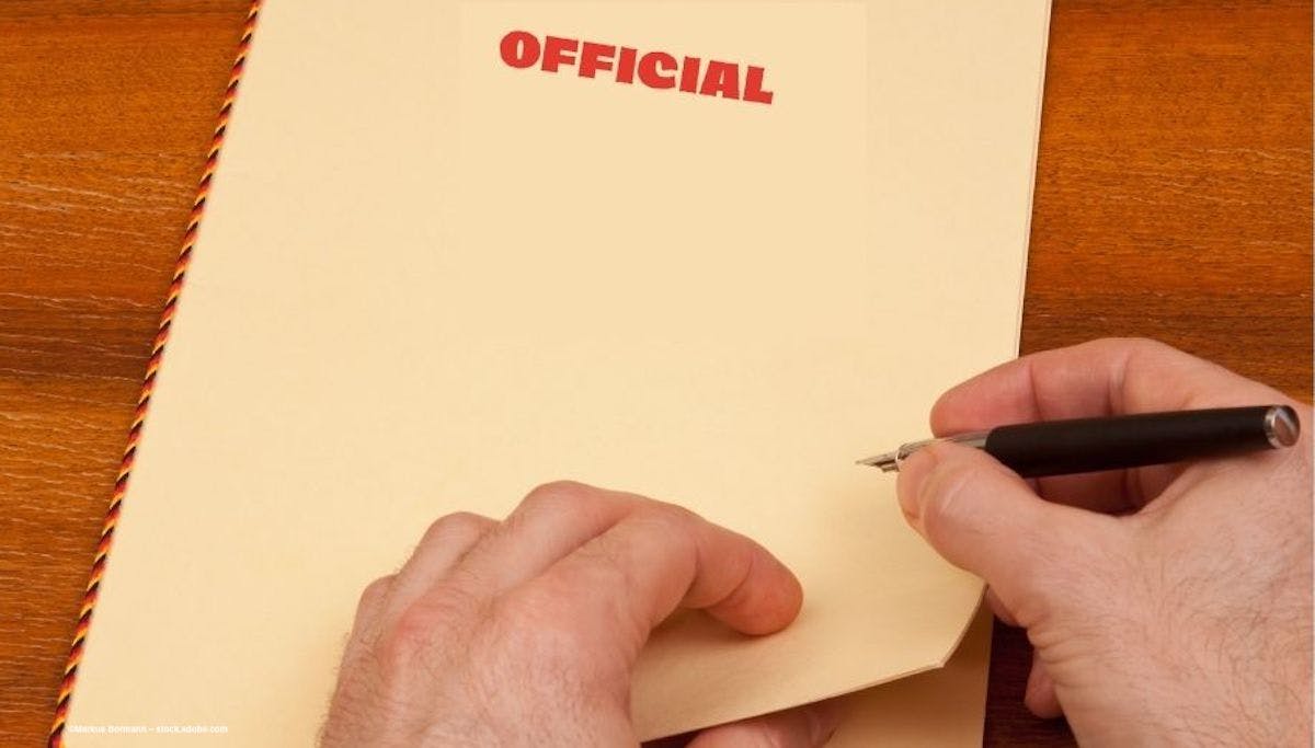 A person at a desk prepares to open a manila file folder with "OFFICIAL" stamped on it. Image credit: ©Markus Bormann – stock.adobe.com