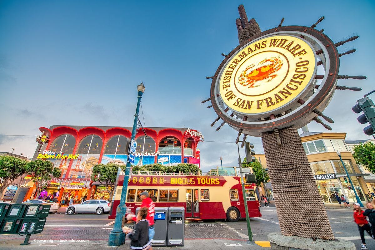 A sign reading "Fisherman's Wharf San Francisco" in front of a bustling tourist area. Image credit: ©jovannig – stock.adobe.com 