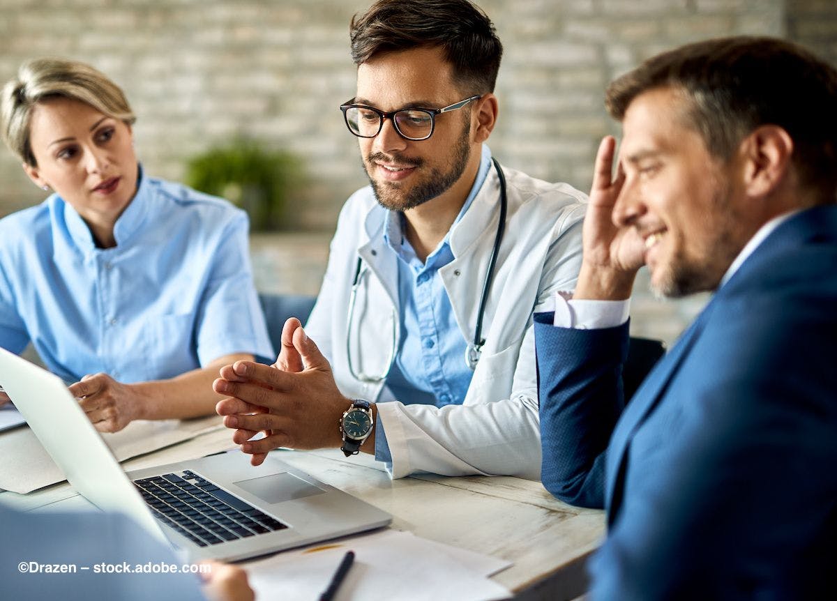 A team of doctors and businesspeople sit at a table, having a discussion. Image credit: ©Drazen – stock.adobe.com