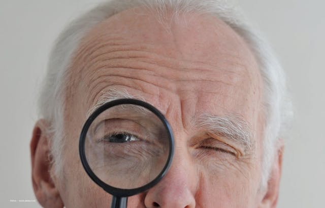 An elderly man holds a magnifying glass close to his eye. Image credit: ©Irina – stock.adobe.com