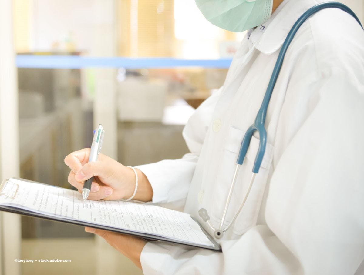A masked physician in white clinical attire writes on a clipboard. Image credit: ©toeytoey – stock.adobe.com