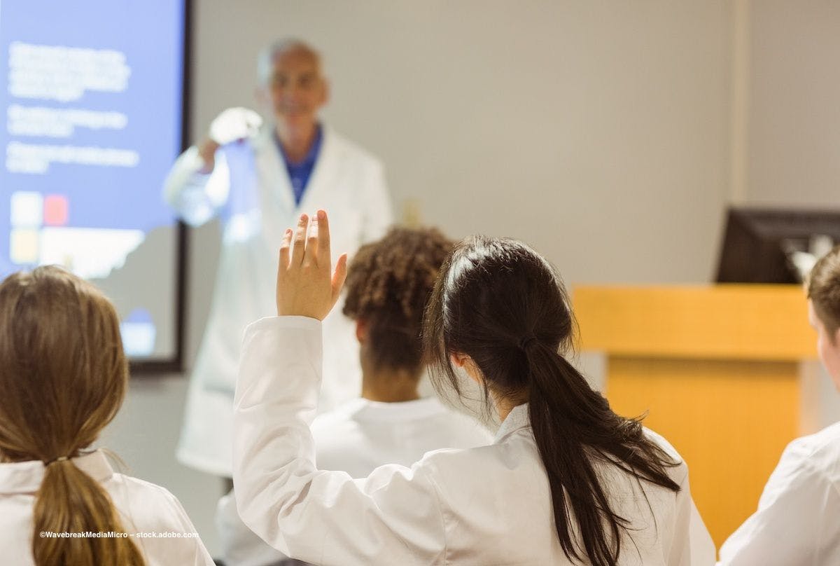 A medical student raises her hand during a lecture as the professor smiles. Image credit: ©WavebreakMediaMicro – stock.adobe.com