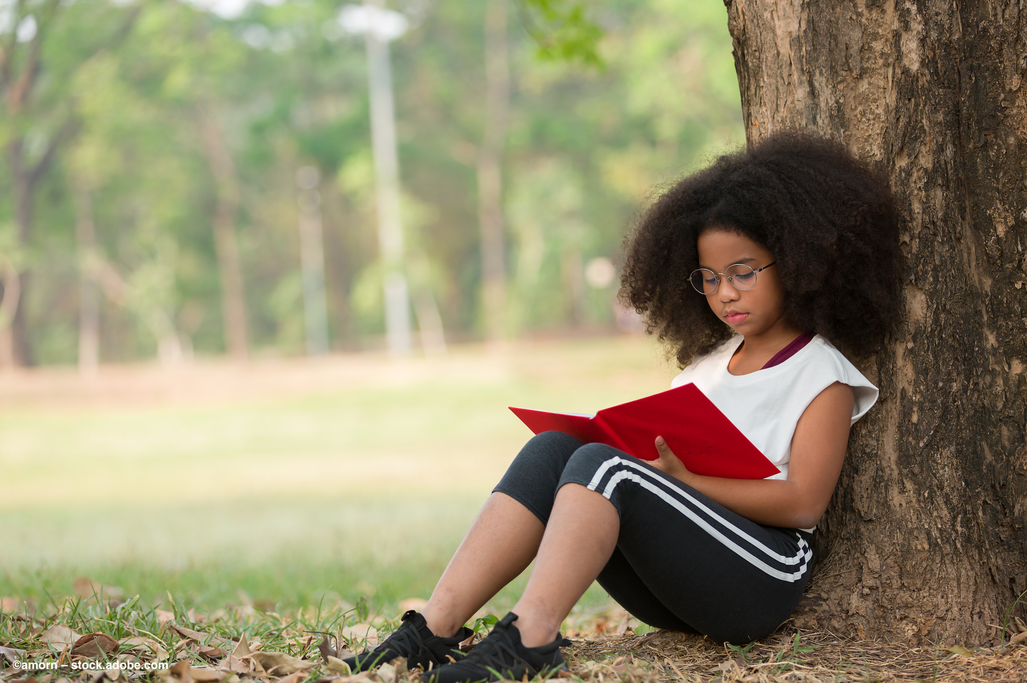 A young girl wearing glasses sits underneath a tree and reads a book. Image Credit: ©amorn – stock.adobe.com