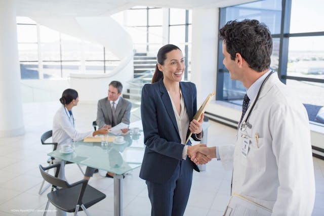 A woman in a business suit shakes hands with a man in a lab coat as their colleagues converse behind them. Image credit: ©Chris Ryan/KOTO – stock.adobe.com